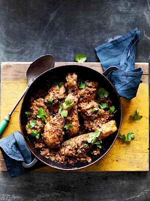 Photograph of Black Chicken Curry with Black Masala Spice Mix in a pan on a table with blue napkins and a yellow board.